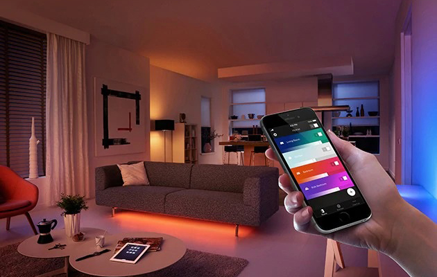 Synkroniser dine lys med solopgang og solnedgang En hånd holder en smartphone foran en stue i farverig belysning.