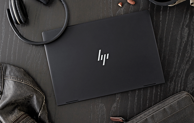 Flot, stilfuldt, praktisk og let design HP Envy x360 på et sort bord omgivet af høretelefoner, en jakke, en taske og peanuts