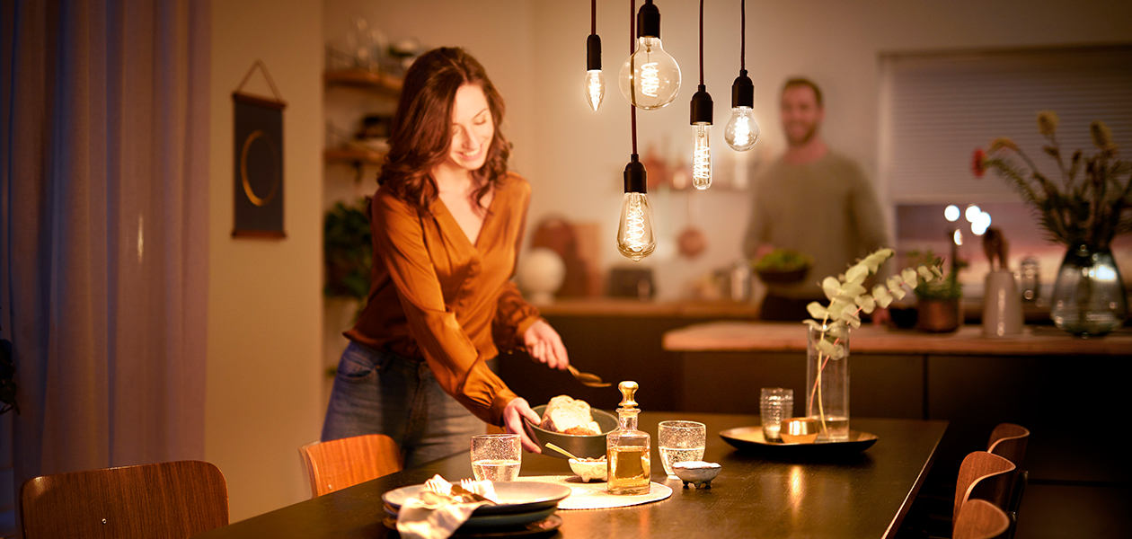 Philips LED Vintage - designad för att synas En leende kvinna som ställer fram mat på bordet, och en man står i bakgrunden.