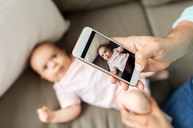 Mand tager billede af baby med sin telefon” title=