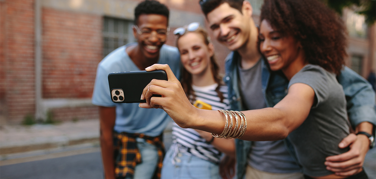 Tech21 – intelligent beskyttelse= Fire venner står til et selfie, en af dem med en mobil med sort Studio Color cover i hånden