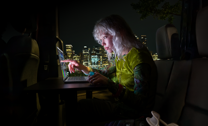 En person sidder på bagsædet af en minibus, den åbne passagerdør viser en naturskøn by ved nattetide, MacBook Pro med PROLED XDR-skærm på et skrivebord, personen oplyses af skærmen, mens vedkommende bruger touchfunktionen til at redigere en video i DaVinci Resolve Studio