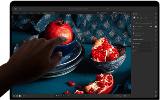 MacBook Pro med PROLED XDR-skærm, der viser et stillbillede af granatæbler, blå keramik og tekstiler, en hånd rækker ud mod skærmen for at interagere med billedet