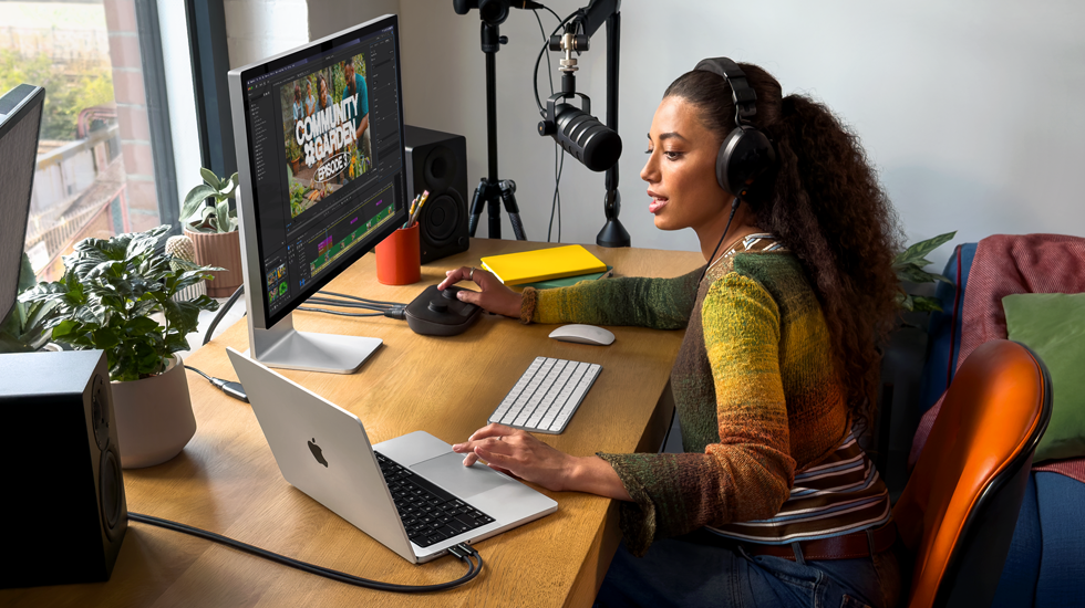 En kvinde arbejder på MacBook Pro hjemme ved sit skrivebord med hovedtelefoner på og en ekstern skærm tilsluttet, mens hun taler i en mikrofon
