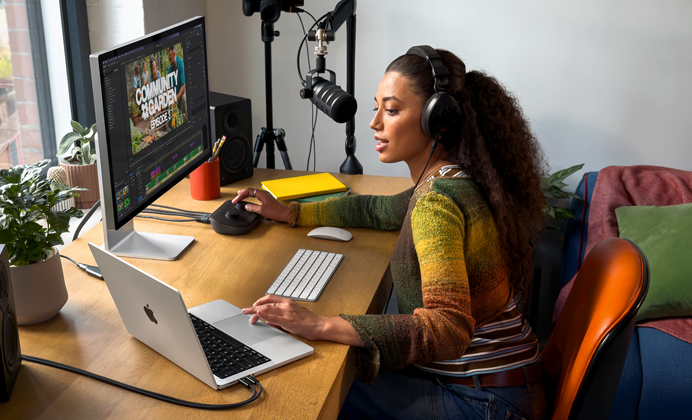 En kvinde arbejder på MacBook Pro hjemme ved sit skrivebord med hovedtelefoner på og en ekstern skærm tilsluttet, mens hun taler i en mikrofon