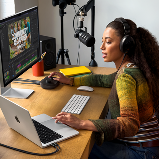 En kvinde arbejder på MacBook Pro hjemme ved sit skrivebord med hovedtelefoner på og en ekstern skærm tilsluttet, mens hun taler i en mikrofon