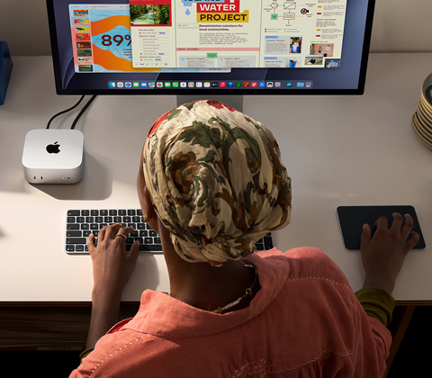 En person sidder ved et skrivebord og skriver med en Mac mini, Studio Display tilsluttet, Magic Keyboard, Magic Trackpad og forskellige ting