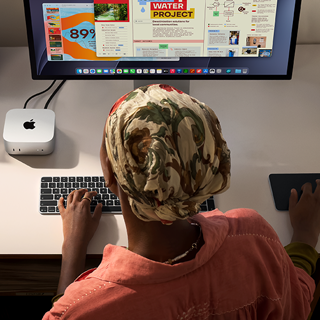 En person sidder ved et skrivebord og skriver med en Mac mini, Studio Display tilsluttet, Magic Keyboard, Magic Trackpad og forskellige ting