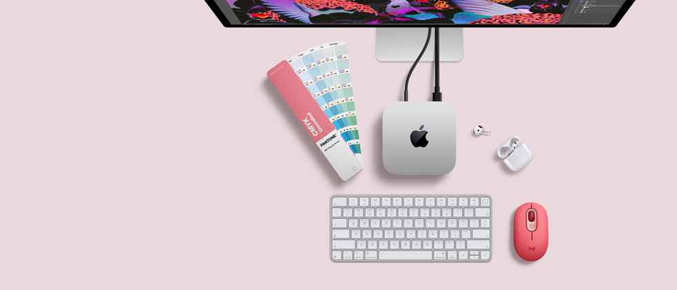 Mac mini i sølvfarvet aluminium vist ovenfra med afrundede hjørner, skinnende sort Apple-logo i midten. Forbundet med et kabel. Studio Display er delvist synligt ovenfra sammen med åbne AirPods 4, Pantone-farvehæfte samt Logitech-tastatur og -mus i rød