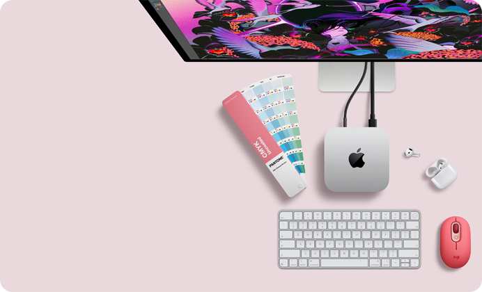 Mac mini i sølvfarvet aluminium vist ovenfra med afrundede hjørner, skinnende sort Apple-logo i midten. Forbundet med et kabel. Studio Display er delvist synligt ovenfra sammen med åbne AirPods 4, Pantone-farvehæfte samt Logitech-tastatur og -mus i rød