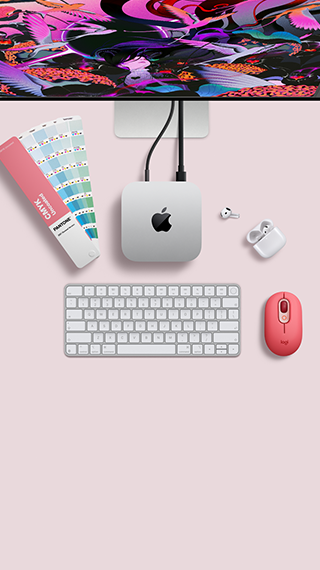 Mac mini i sølvfarvet aluminium vist ovenfra med afrundede hjørner, skinnende sort Apple-logo i midten. Forbundet med et kabel. Studio Display er delvist synligt ovenfra sammen med åbne AirPods 4, Pantone-farvehæfte samt Logitech-tastatur og -mus i rød