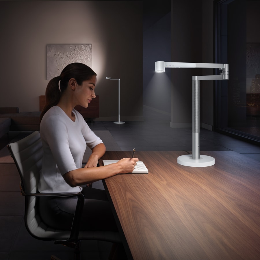 Kvinde, der sidder ved et bord og skriver, med en Dyson-lampe foran sig