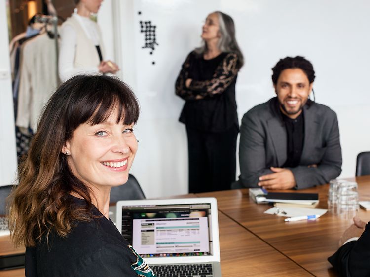En kvinde og to mænd sidder smilende rundt et bord i et mødelokale og arbejder på en bærbar computer