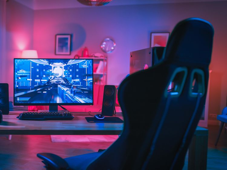 Gamingopsætning med skærm, tastatur og højttalere på et bord, mens en gaming-stoll ses foran det.