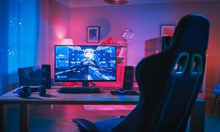 Gamingopsætning med skærm, tastatur og højttalere på et bord, mens en gaming-stoll ses foran det.
