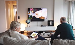 Ældre kvinde sidder i en sofa med hendes tablet, mens hendes mand sidder og ser TV