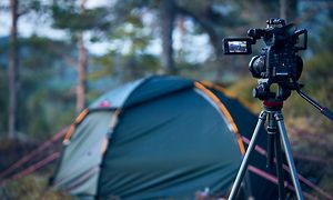 Kamera på stativ stående i en skov, der peger mod et telt