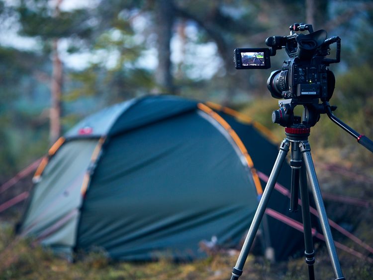 Kamera på stativ stående i en skov, der peger mod et telt