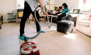 Mand støvsuger, mens kone og børn sidder i sofaen bag ham