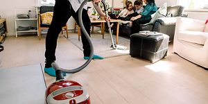 Mand støvsuger, mens kone og børn sidder i sofaen bag ham
