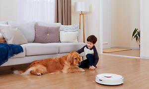 Dreng og hund sidder på et trægulv ved siden af en hvid robotstøvsuger