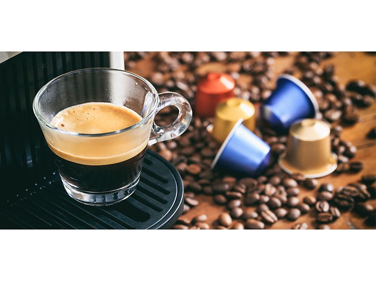En kop espresso fra en kapselmaskine med kapsler og kaffebønner på disken