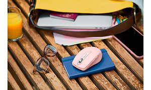 Logitech mus på en notesbog ved siden af briller og taske med papirer på et havebord.
