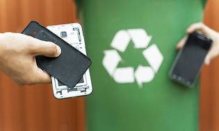 To hænder viser gamle mobiltelefoner foran et grønt genbrugsskilt