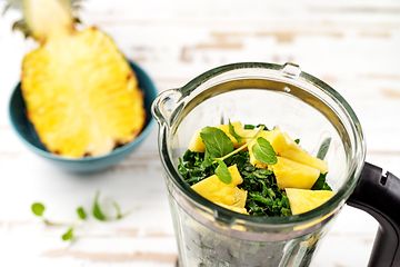 En halv ananas i en blå skål ved siden af en blender med grønt og ananas