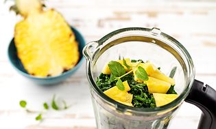 En halv ananas i en blå skål ved siden af en blender med grønt og ananas