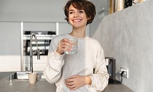 Smilende kvinde står i et køkken og nyder et glas vand