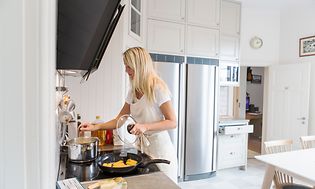 Kvinde laver mad i køkken med et køleskab og et fryseskab i baggrunden