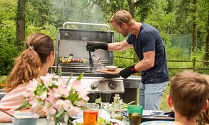 En mand griller på en gasgrill mens familien ser til