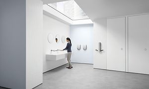 Kvinde vasker hænder på et offentligt toilet med Dyson AirBlade-tørretumblere ved siden af sig