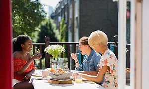 Tre kvinder sidder rundt et bord på en altan og smiler mens de spiser
