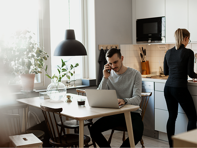 En mand, der arbejder på sin bærbare computer og taler på sin smartphone i et køkken