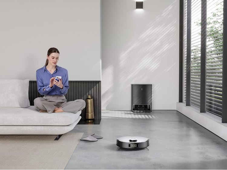 SDA - Ecovacs - Deebot X1 på gulvet hvor den gør rent og der sidder en kvinde i en sofa ved siden af