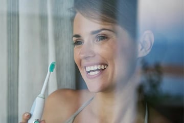 En kvinde, der smiler i et vindue, mens hun holder en elektrisk tandbørste