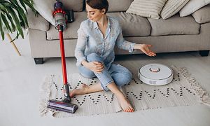 En kvinde sidder på gulvet mellem en dyson trådløs støvsuger og en hvid robotstøvsuger