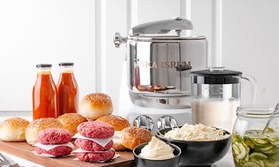 Hamburgerkød, boller og aioli og glas med syltede agurker, og en Ankarsrum køkkenmaskine på et køkkenbord