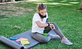 En kvinde tjekker sit smartwatch, mens hun sidder på en yogamåtte udendørs 