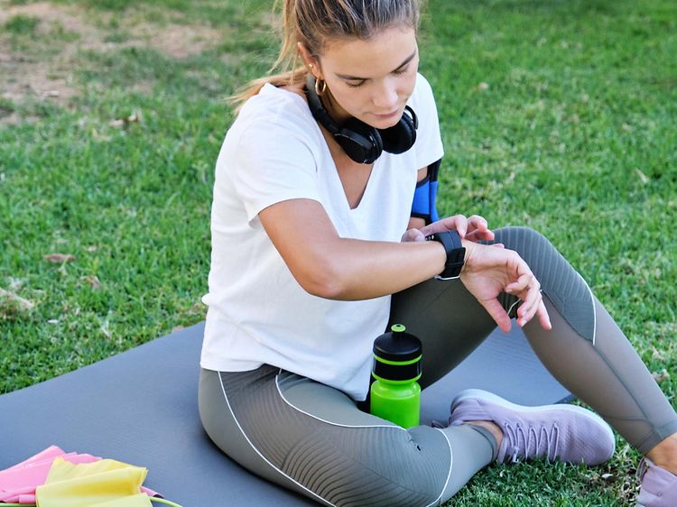 En kvinde tjekker sit smartwatch, mens hun sidder på en yogamåtte udendørs 