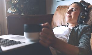 Kvinde med hovedtelefoner, bærbar computer og kaffekop ligger i sofa
