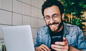 En smilende mand med en smartphone foran en bærbar computer