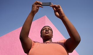Kvinde står med Pixel 6a i hænderne med lyserød bygning og himmel bagved