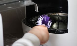 En person rengør airfryer med en lilla opvaskebørste