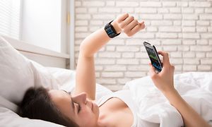 Telecom - Workout - En kvinde der ligger i sengen og kigger på sin pulsmåler med en telefon i hånden