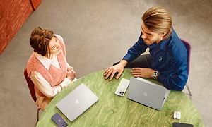 Apple - Økosystem - Mandlig Elkjop-medarbejder taler med en kvinde ved et bord med Apple-produkter