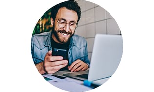 Smilende mand der sidder foran en laptop med en smartphone i hænderne