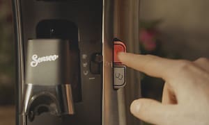 Nærbillede af en hånd, der trykker på tænd-knappen på en Senseo Select-kaffemaskine